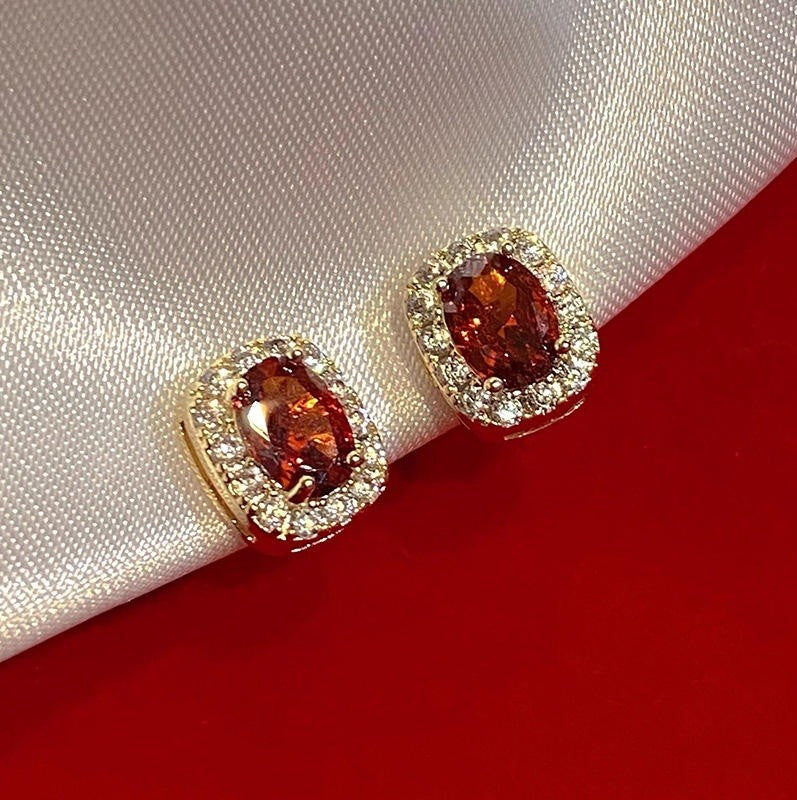 A pair of gold-toned oval stud earrings with marquise-cut orange-red center stones and clear halo stones, resting on a white satin fold over a red background.