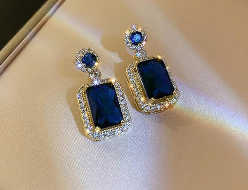 A pair of gold drop earrings featuring large rectangular dark blue gemstones surrounded by small clear stones, with smaller round blue stones above, displayed on a beige satin surface.