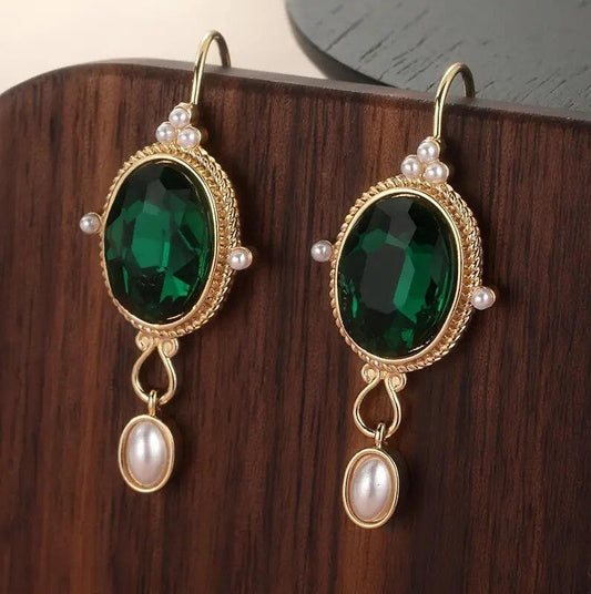 A pair of gold-tone dangle earrings with large oval emerald green crystals, accented with small white faux pearls, displayed on a dark wood block.