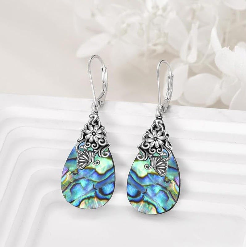 A pair of silver drop earrings with ornate filigree tops and teardrop-shaped abalone shell pendants showing iridescent blue, green, and purple tones, displayed on a white surface with soft white flowers in the background.