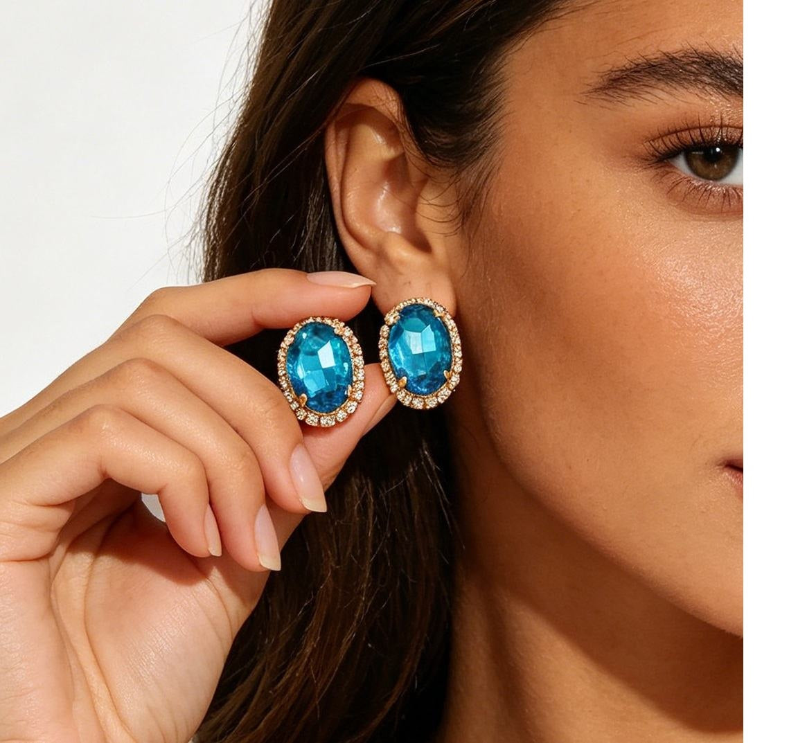 A close-up lifestyle shot of a model wearing oval blue gemstone stud earrings with gold-toned halo settings and small clear accent stones, while her other hand gently holds the matching earring against a clean white background.