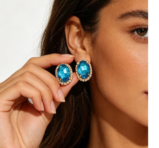 A close-up lifestyle shot of a model wearing oval blue gemstone stud earrings with gold-toned halo settings and small clear accent stones, while her other hand gently holds the matching earring against a clean white background.