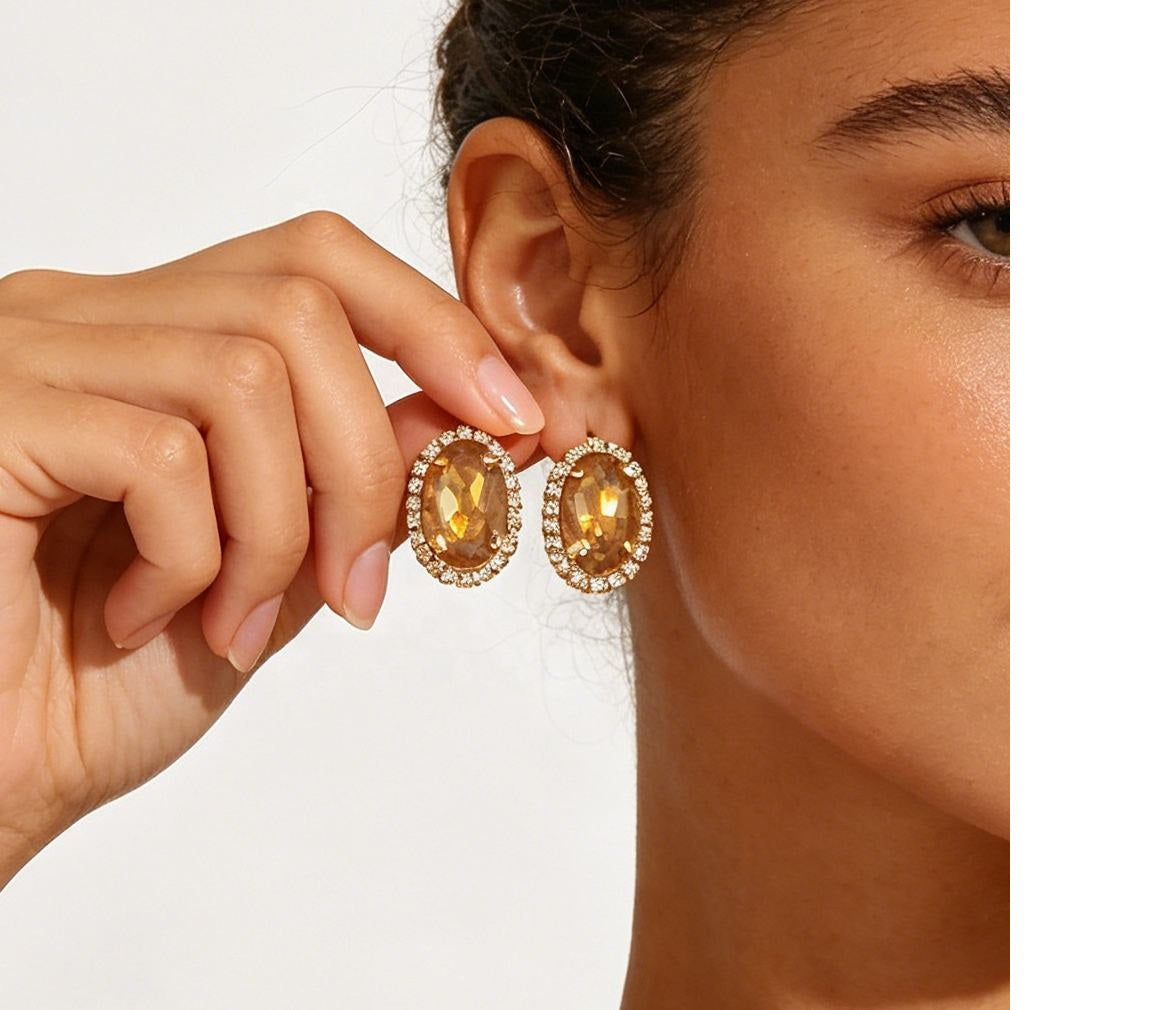 A close-up of a model wearing oval drop earrings with large amber-yellow center stones surrounded by small clear stones in a gold-tone setting, with one earring being held near the ear against a plain light background.