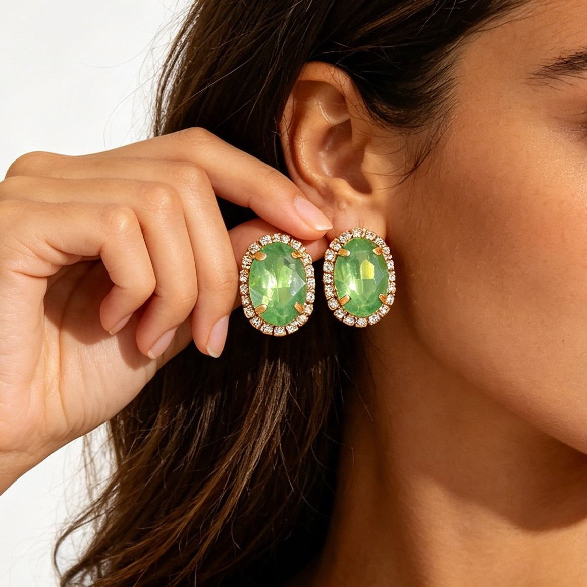 Close-up of a woman wearing oval green gemstone earrings with gold-tone halo settings and small clear stones, with one earring being adjusted by a hand.