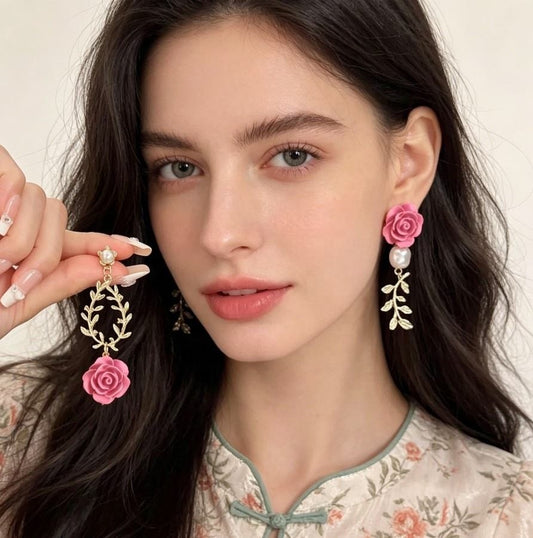 A close-up of a woman wearing dangling earrings with pink roses, gold leaf-like branches, and pearl accents, while holding one matching earring in her hand.