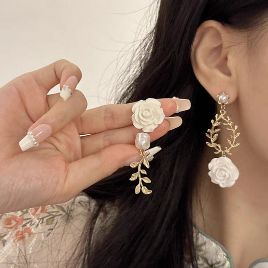 A close-up of a person wearing a dangling earring while holding the matching earring in their hand. The earring features a white rose, a pearl, and a gold leaf vine design.