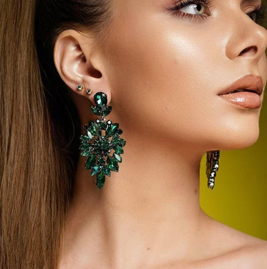 A close-up of a woman's ear wearing a large, elaborate green gemstone dangle earring with multiple cascading leaf-like clusters. The earring features deep emerald green stones with dark metal settings.