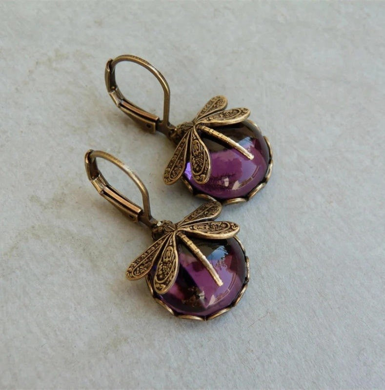 A pair of dangling earrings with antique bronze dragonfly charms and oval purple cabochon stones, displayed on a light textured surface.