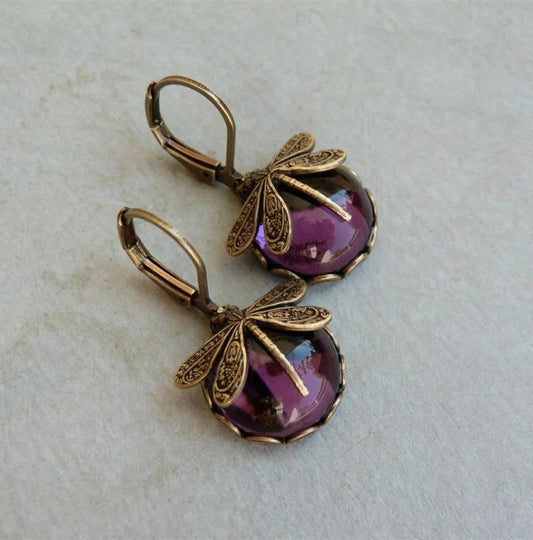 A pair of dangling earrings with antique bronze dragonfly charms and oval purple cabochon stones, displayed on a light textured surface.