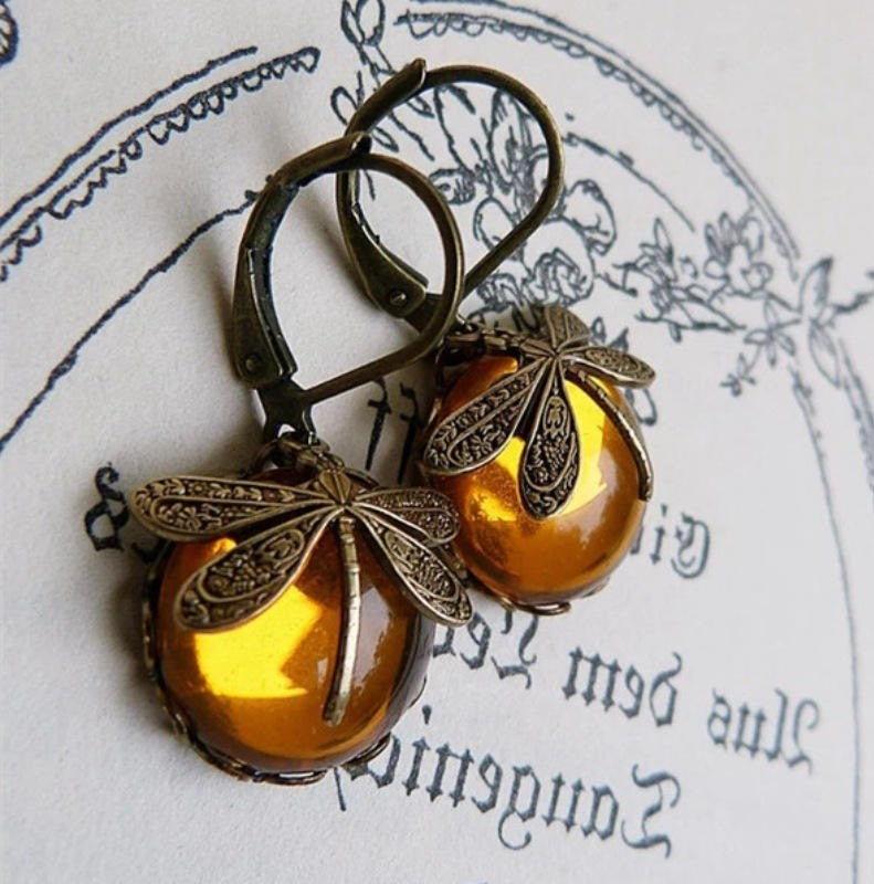 A pair of antique-style dragonfly earrings with amber-colored glass beads and ornate filigree metal wings, displayed on a piece of paper with cursive writing.