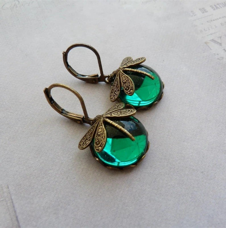 A pair of dangling earrings with antique bronze dragonfly charms and faceted green glass beads, displayed on a light gray textured surface.