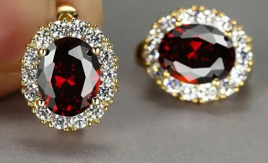 A pair of oval halo earrings featuring deep red faceted center stones surrounded by clear pavé-set stones in yellow gold settings, with one earring held against a finger and the other resting on a neutral gray surface.