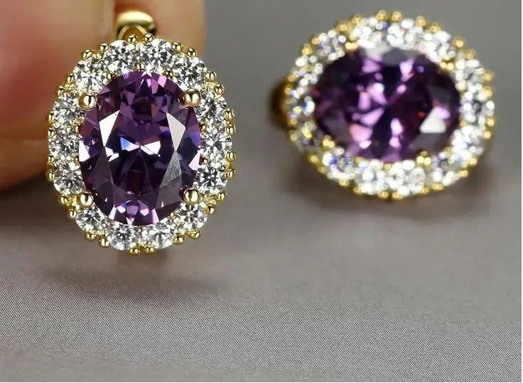A pair of oval halo earrings featuring a large faceted purple gemstone surrounded by a halo of small clear stones in yellow gold settings, shown on a neutral gray background.