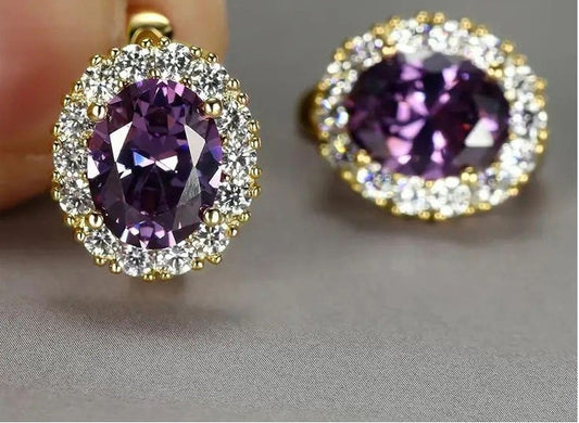 A pair of oval halo earrings featuring a large faceted purple gemstone surrounded by a halo of small clear stones in yellow gold settings, shown on a neutral gray background.