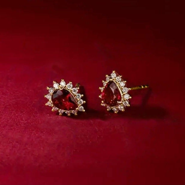 A pair of gold stud earrings featuring a central teardrop-shaped deep red gemstone surrounded by a halo of small clear stones, displayed on a textured dark red background.