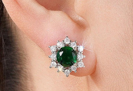 A close-up of a person's ear wearing a silver-toned stud earring with a round green emerald-cut center stone surrounded by a halo of small clear stones.