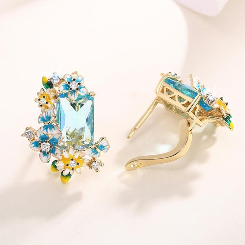 A pair of gold hoop earrings with a large rectangular light blue gemstone and colorful floral enamel details in blue, yellow, and white, displayed on a clean white background.