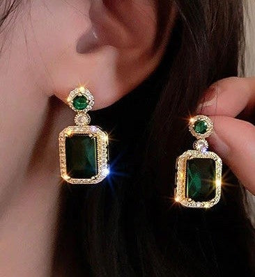 A close-up of a person wearing and holding a pair of gold drop earrings with large rectangular green gemstones and small clear stones around the settings.