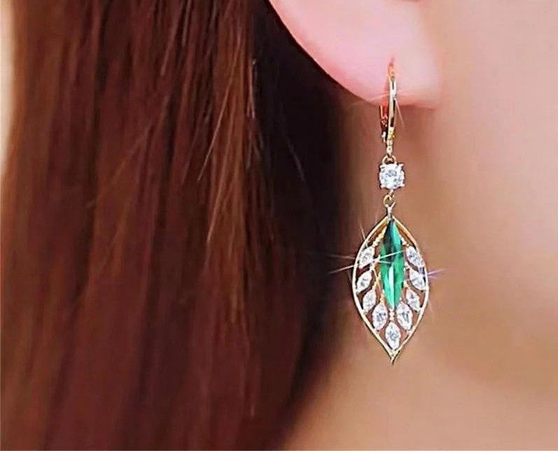 A gold drop earring with a marquise-cut green center stone and clear accent stones, shown worn on a person's ear with brown hair visible.