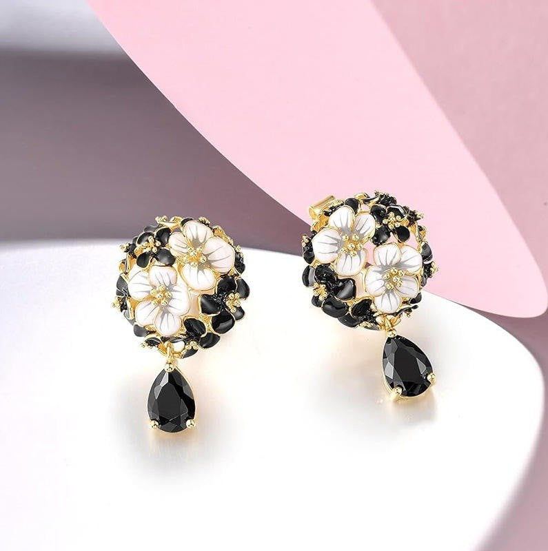 A pair of gold-tone floral drop earrings with white and black flower details, displayed on a white surface with a pink and gray background.
