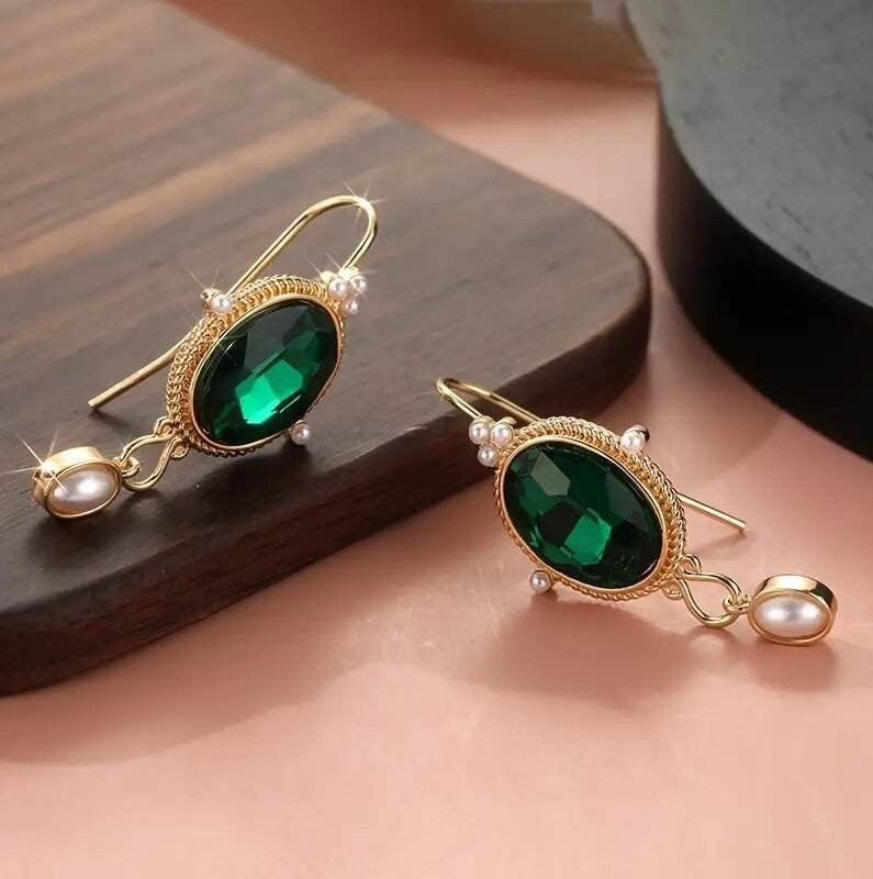 A pair of gold drop earrings with oval green faceted gemstones, small pearl-like beads, and dangling pearl drops, displayed on a dark wooden block against a soft pink background.