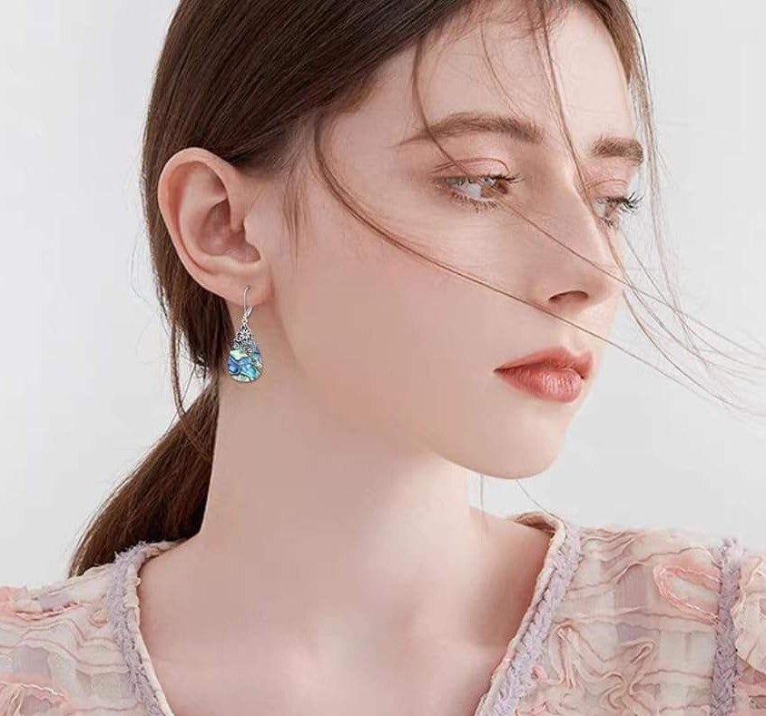 A close-up of a woman wearing a dangling earring with a light blue faceted gemstone and silver-toned metal, against a soft light gray background.