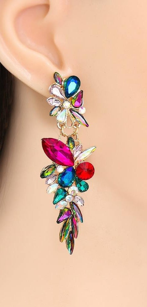 A close-up of a person's ear wearing a dangling statement earring with colorful rhinestones and leaf-like floral details in blue, pink, red, green, and purple on a gold-tone setting.