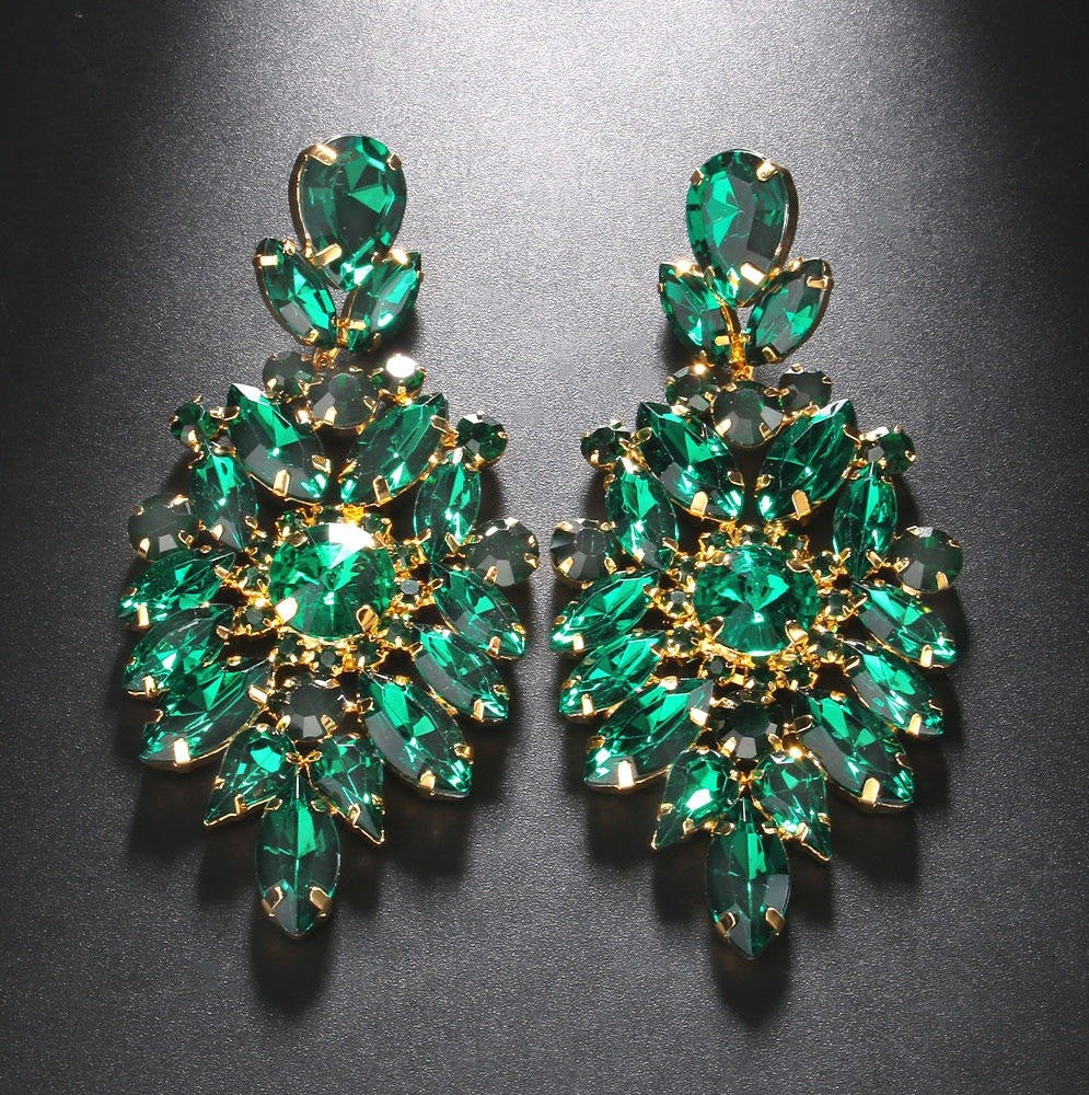 A pair of large gold-tone statement earrings adorned with multiple emerald green marquise and oval rhinestones, displayed on a dark textured background.
