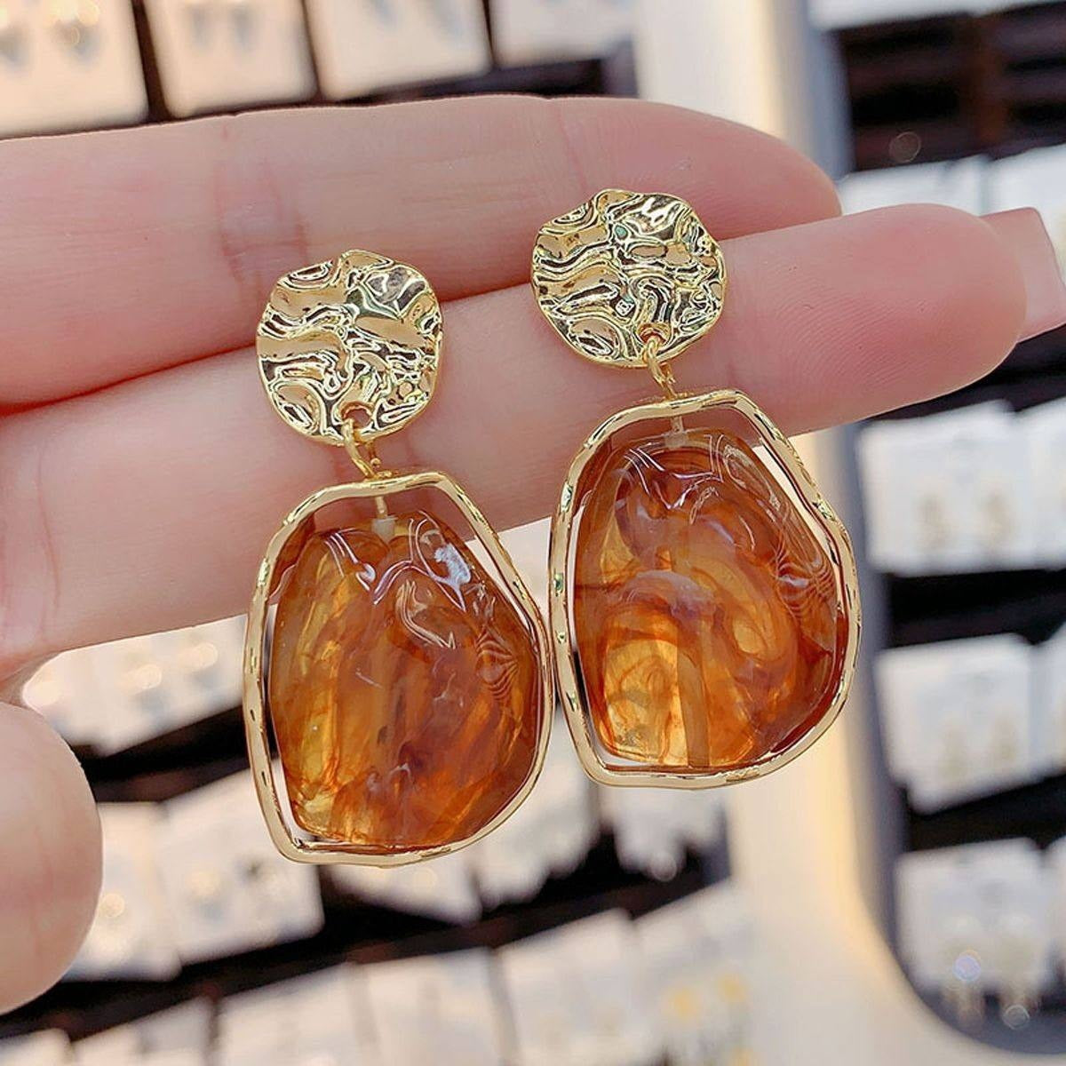 Gold earrings with amber-colored stones held between fingers against a blurred background