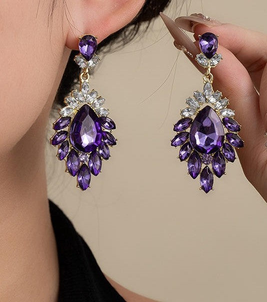 A pair of gold-tone drop earrings with large faceted purple teardrop gemstones and smaller clear rhinestone accents, shown worn on one ear and held by a hand against a plain beige background.