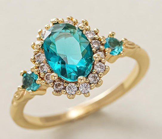 A yellow gold ring featuring a large oval-cut turquoise gemstone in a halo setting with small clear stones, accented by three smaller round turquoise gemstones on the band, set against a light beige background.