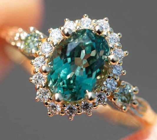 An oval-cut emerald-cut green gemstone ring set in yellow gold with a halo of small clear round-cut stones, shown close-up on a blurred neutral background.