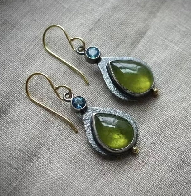 A pair of handmade teardrop dangle earrings featuring olive green gemstone drops in textured silver settings with small blue round stones and gold-tone fishhook wires, displayed on a neutral linen fabric background.
