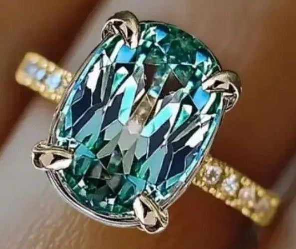 A close-up of a ring worn on a finger, featuring a large oval-cut green gemstone in a silver-toned setting with a yellow-gold band accented by small clear stones.