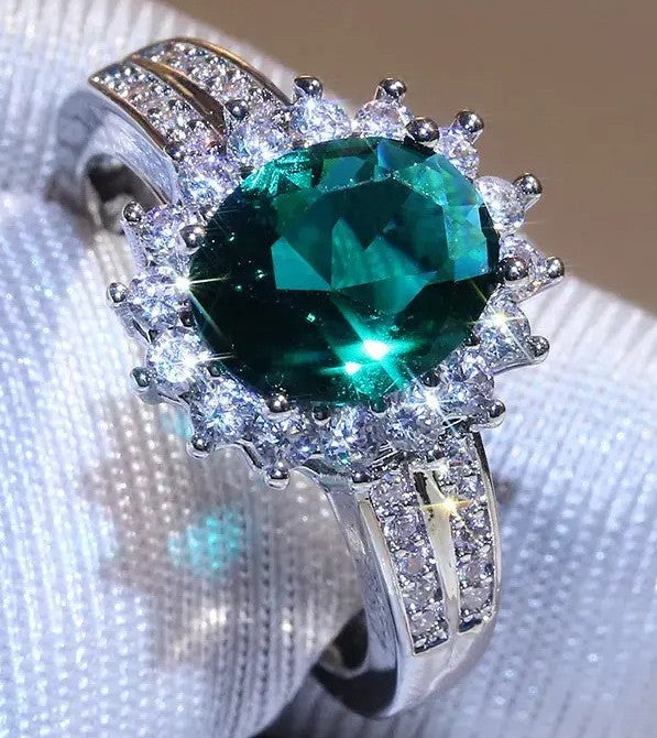 A silver-tone halo ring featuring a large oval-cut green emerald gemstone surrounded by small clear stones, displayed on a white textured fabric surface.