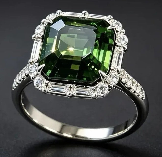 A silver-toned ring featuring a large, cushion-cut green gemstone surrounded by a halo of small clear stones, with baguette and round-cut accents on the band, set against a dark background.