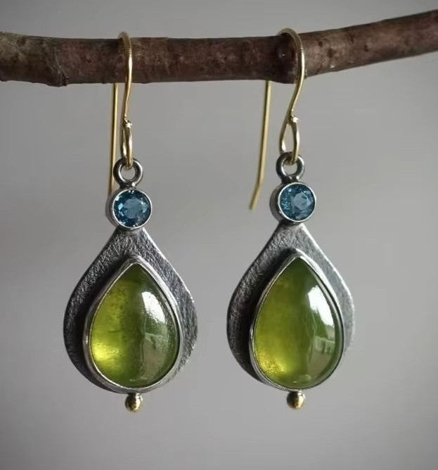A pair of handmade teardrop dangle earrings hanging from a rustic brown branch. The earrings feature a textured silver-toned metal frame, a small round blue gemstone, and a larger polished olive-green teardrop stone.