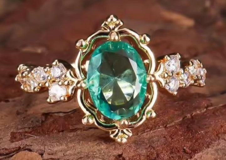 A gold ring featuring a large oval green gemstone in the center, flanked by small clear stones on the band, set against a textured brown background.
