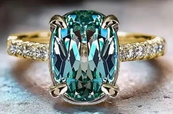 A yellow gold ring featuring a large oval-cut emerald-cut green gemstone in a four-prong setting, with small clear accent stones along the shoulders, shown on a textured gray surface.