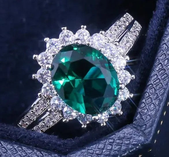 A silver-tone halo ring featuring a large oval-cut green emerald gemstone surrounded by clear stones, displayed on a dark velvet ring box.