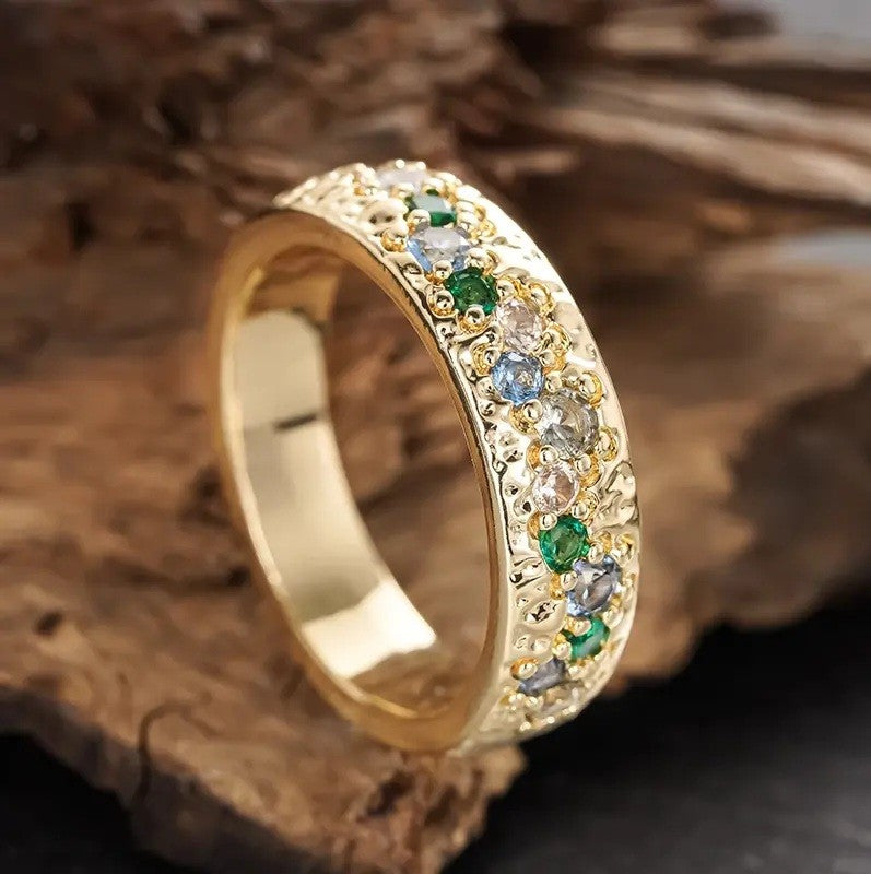 A gold band ring decorated with a row of round-cut clear, light blue, and green gemstones, displayed on a piece of rustic wood.