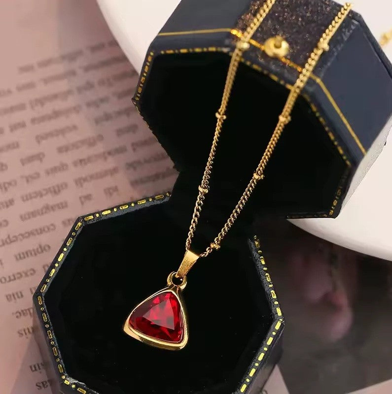 A gold chain necklace with a triangular red gemstone pendant, displayed inside an open black jewelry box.