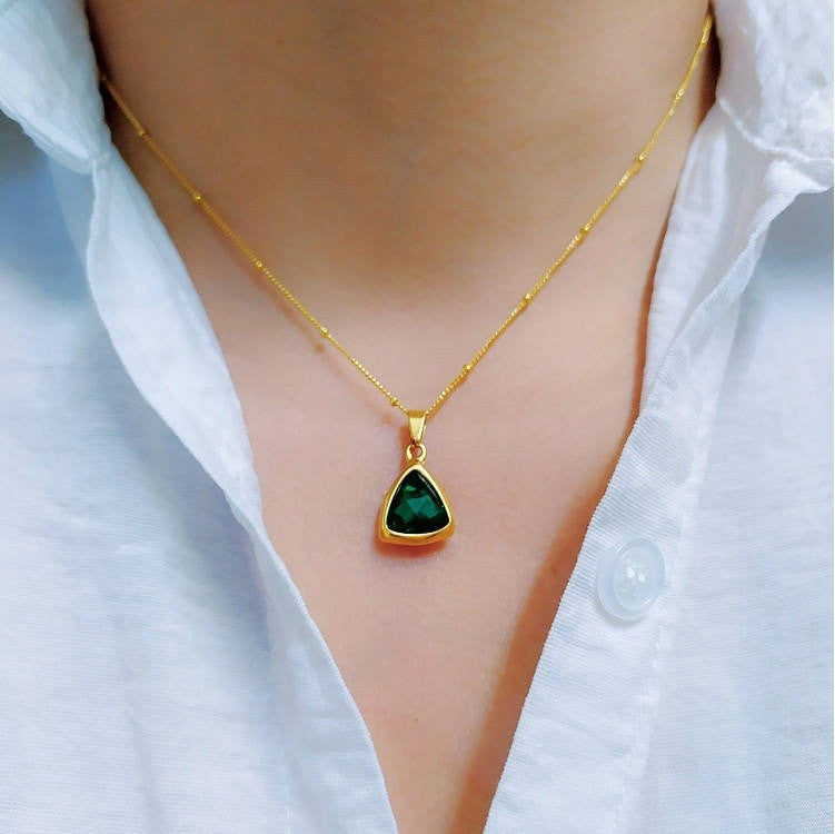 A gold necklace with a triangular pendant featuring a green stone, worn on a person's neck over a white collared shirt.