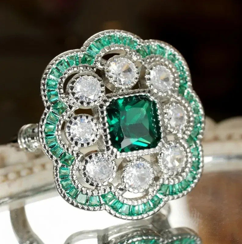 A close-up of an ornate silver-toned ring featuring a central emerald-cut green gemstone surrounded by round clear stones and a halo of smaller green stones, shown against a dark blurred background.