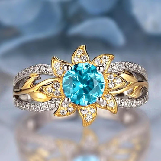 A floral sunburst ring featuring a large round-cut light blue center stone surrounded by gold-toned leaf-like petals, with a silver-toned band paved with small clear stones.