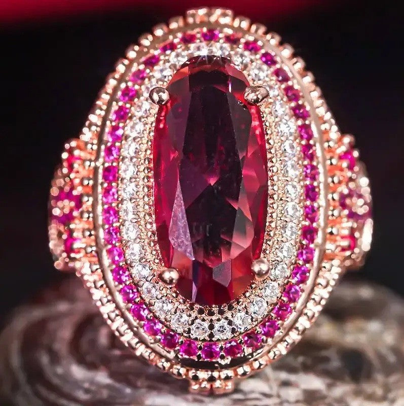 An oval ring featuring a large faceted deep red ruby center stone, surrounded by a double halo of small clear pavé diamonds and a border of round pinkish-red rubies, set in rose gold-toned metal.