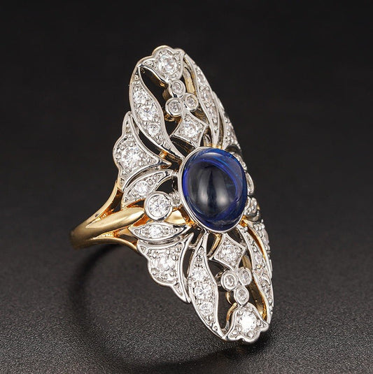 An ornate Art Deco style ring featuring a central oval blue sapphire surrounded by pavé-set clear stones on a yellow gold and white gold setting, photographed on a dark background.