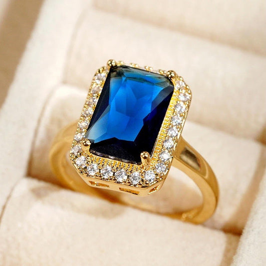 A gold ring featuring a rectangular blue gemstone surrounded by a halo of small clear stones, displayed on a cream-colored ring box.