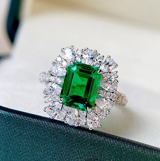 A silver ring featuring a large emerald-cut green gemstone surrounded by a halo of clear round-cut stones, displayed on a dark green satin ribbon over a light cream background.
