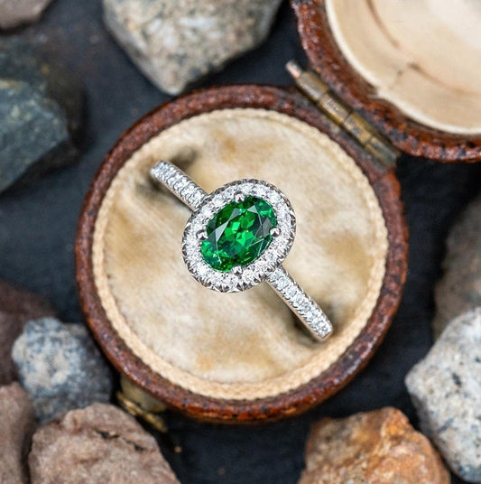 A silver ring with a round green emerald-cut gemstone and a halo of small clear stones, displayed in an open brown ring box on a dark surface with rocks around it.